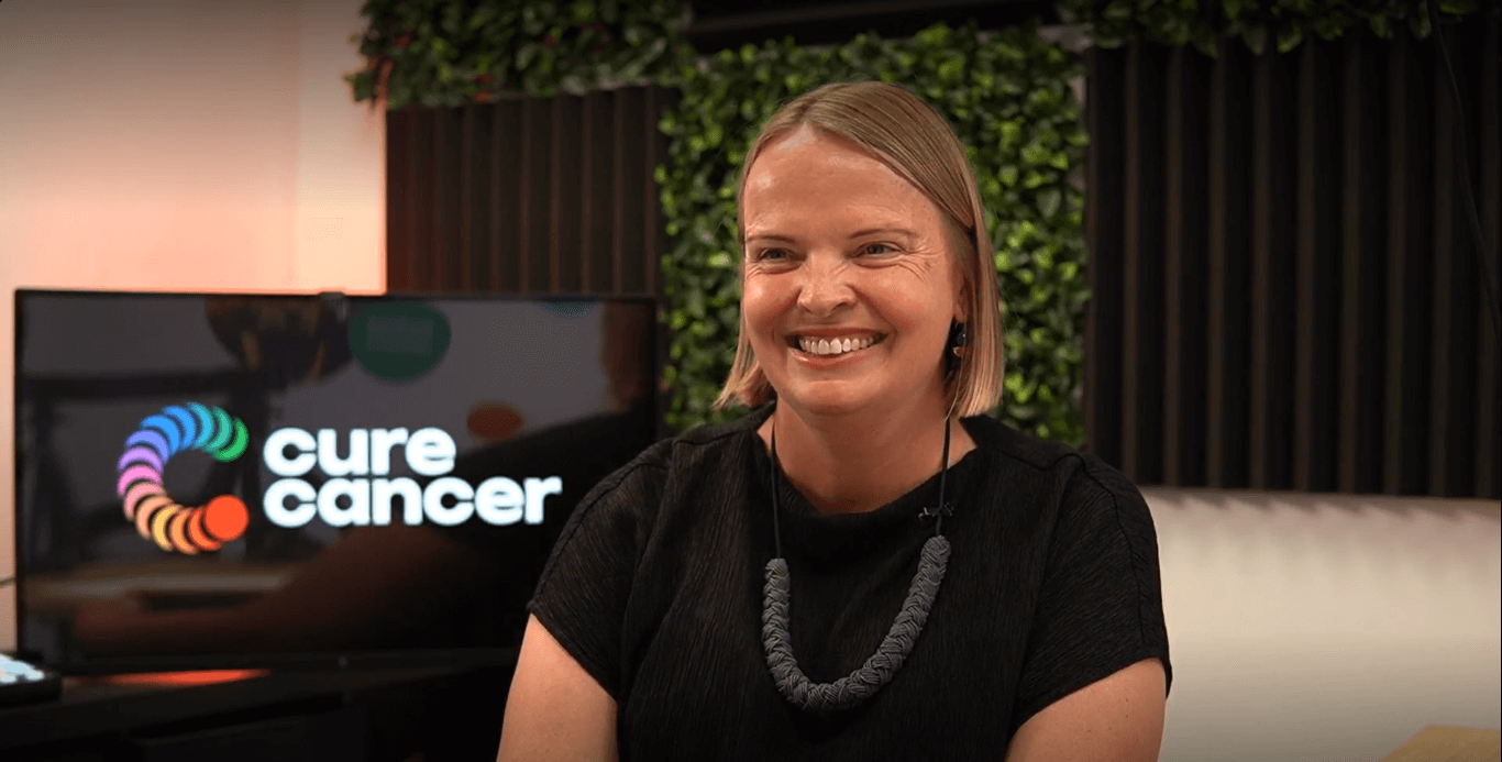A/Prof Caroline Ford smiles while seated in the Cure Cancer office. She is wearing a black shirt and a necklace.