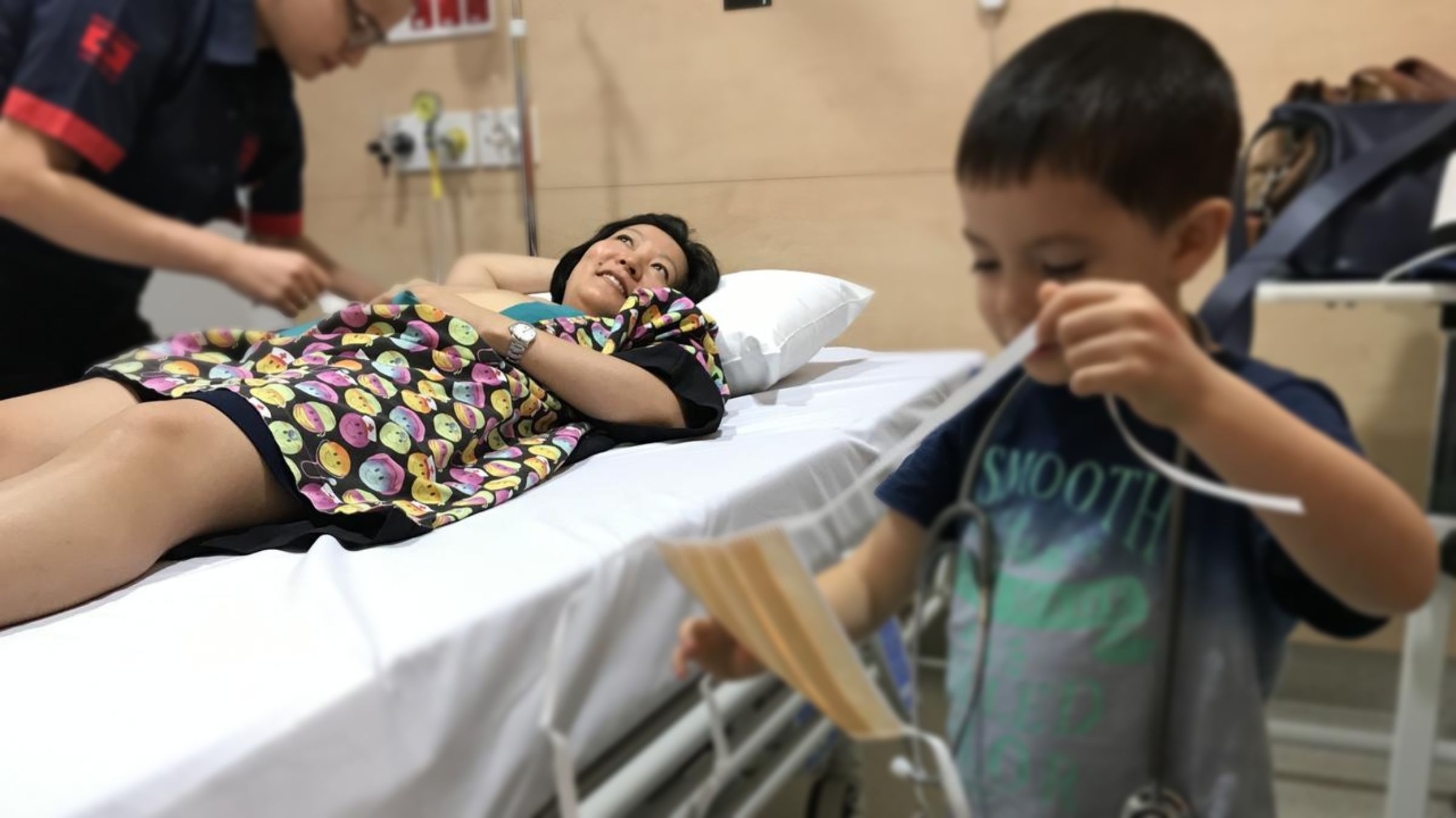 Katrina Lau Hammond lies on a hospital bed as a nurse administers her cancer treatment. Her son is playing off to the side.