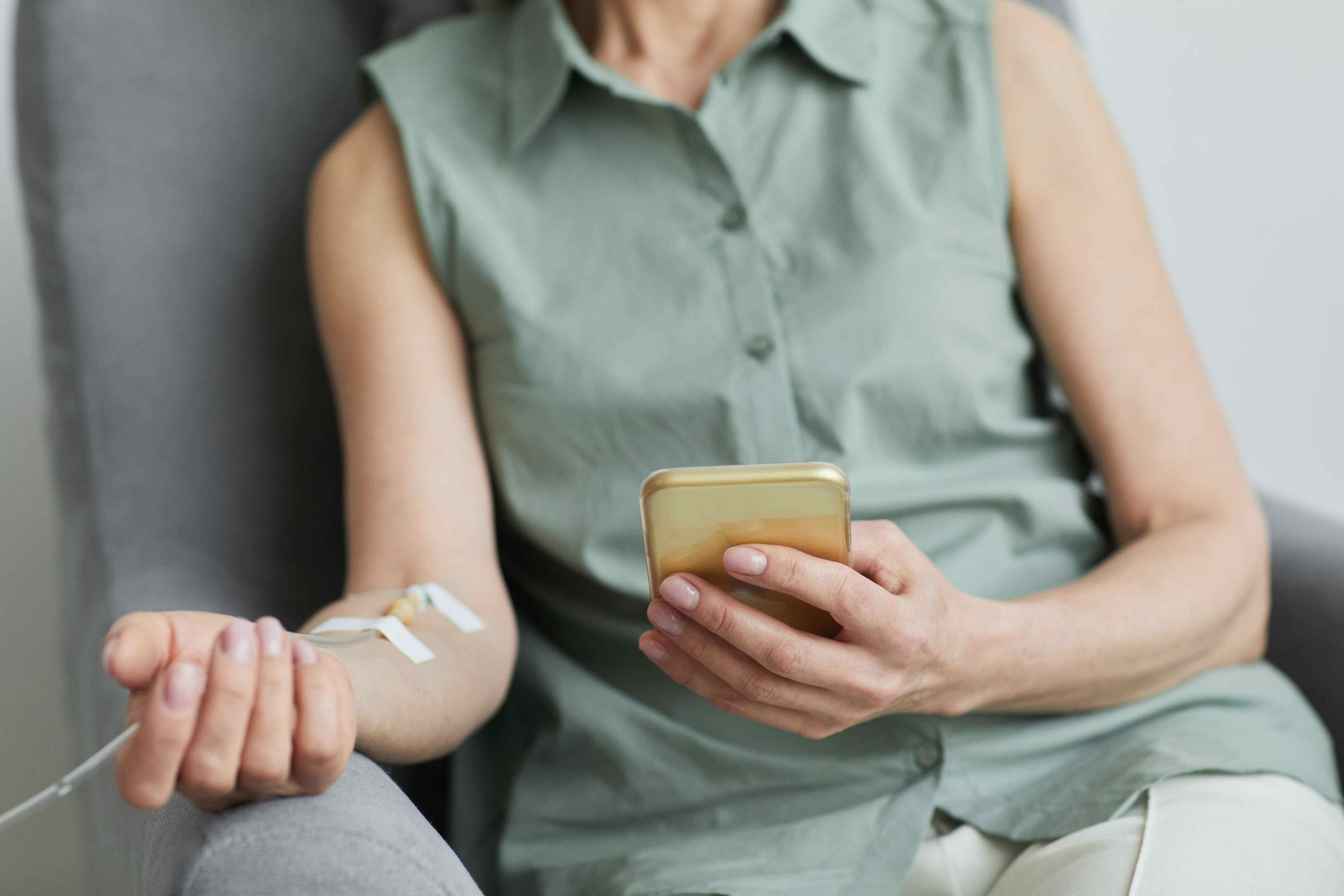 A patient wearing a sage sleeveless top receives a transfusion. They are on their phone.