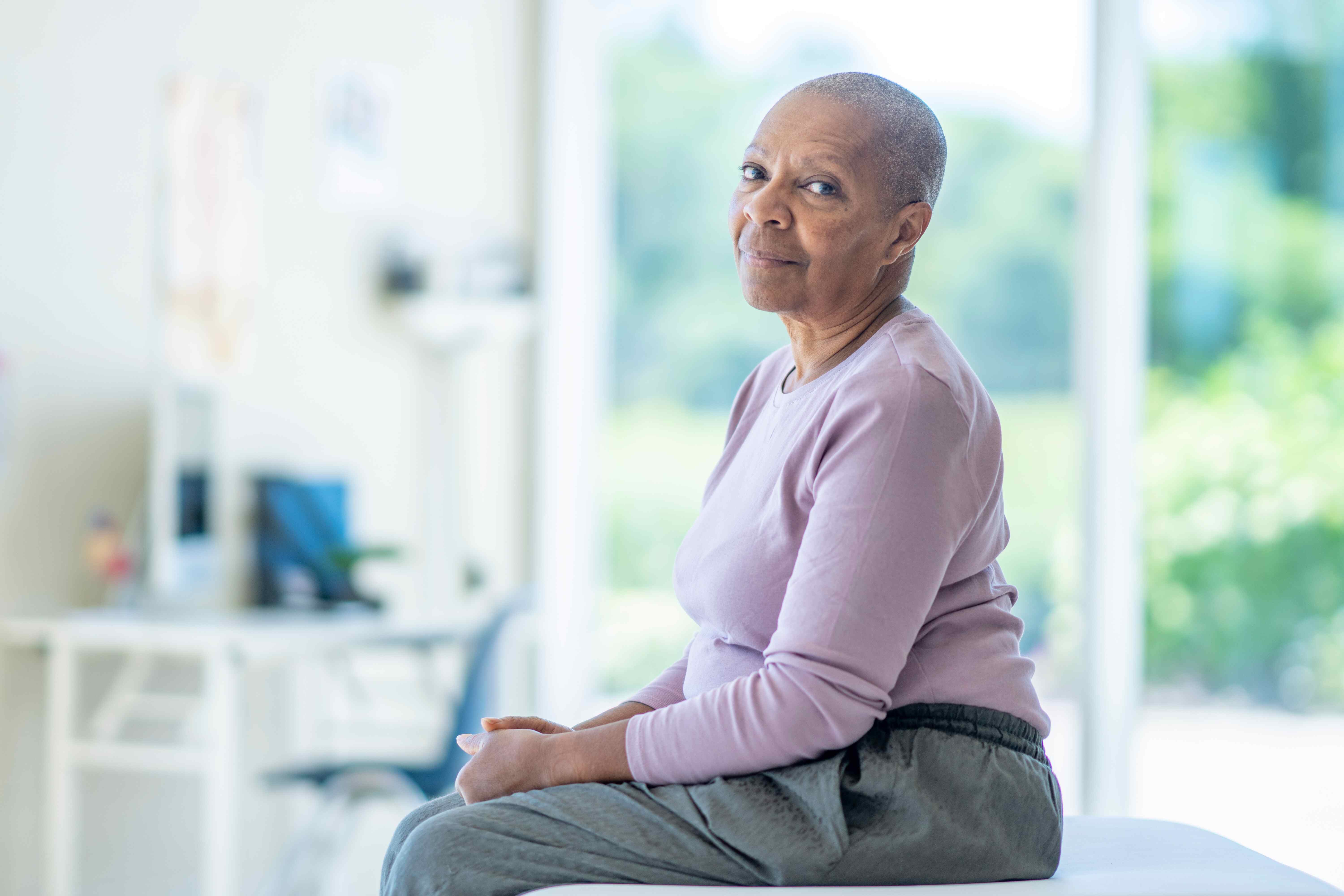 A cancer patient with a shaved head is sitting down. They are wearing a lilac shirt and grey trousers.